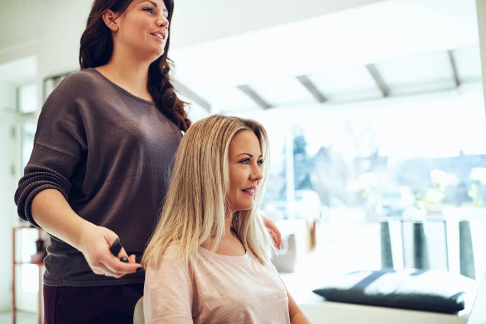 Smiling young blonde woman sitting in a salon chair getting her hair styled during an appointment with her hairdresser
